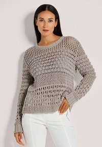 Hellgrauer Strickpullover mit offenem Häkelmuster, langen Ärmeln und gerippten Bündchen. Kombiniert mit weißen Hosen.