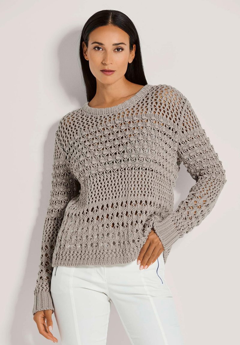 Hellgrauer Strickpullover mit offenem Häkelmuster, langen Ärmeln und gerippten Bündchen. Kombiniert mit weißen Hosen.