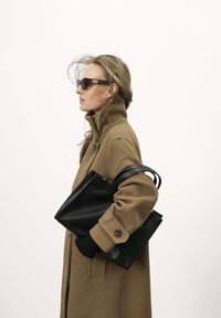Manteau marron avec un col relevé, sac fourre-tout en cuir noir et gants noirs. Lunettes de soleil caractéristiques avec un détail de logo. Fond clair.