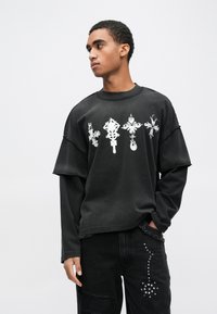 Fekete túlságosan nagy pulóver fehér grafikai nyomatokkal, kerek nyakkivágással és lezuhant vállakkal, párosítva szakadt fekete farmerrel, mely ezüst szegecsekkel díszített.