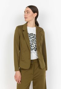 Olivově zelený upravený blazer s klopami, se dvěma knoflíky a hladkým povrchem, nošený přes bílou košili se černo-zeleným leopardím potiskem.