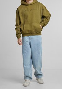 Olivově zelená mikina s kapucí a přední kapsou, s nápisovým logem. Kombinováno se světle modrými volnými džíny a bílými teniskami.