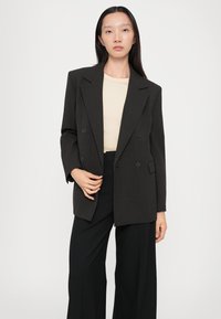 Blazer doppiopetto in tessuto marrone scuro, con revers a punta, due bottoni, tasche frontali, abbinato a una blusa beige chiaro e pantaloni neri.