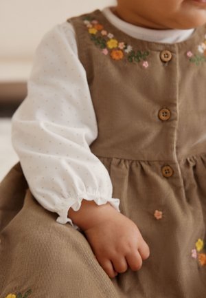 Småbarn iført en brun manchesterkjole med blomstermønsterbroderi og træknapper, over en hvid langærmet skjorte med små prikker.