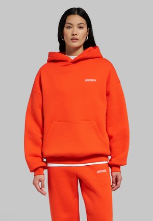 Femme aux cheveux noirs et raides portant un sweat à capuche orange vif et un pantalon assorti avec le texte blanc "WRSTBHVR" sur la poitrine et la cuisse, fond gris.