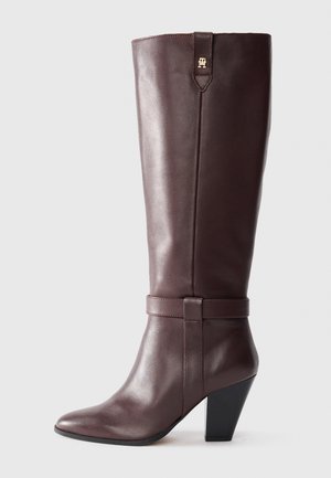 Bota de cuero marrón hasta la rodilla con punta afilada, tacón cónico y un detalle decorativo de correa en el tobillo. Acabado suave y brillante.