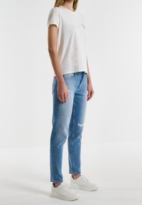 Camiseta blanca de manga corta con un pequeño gráfico, combinada con jeans rasgados de color azul claro. El modelo lleva zapatillas blancas.