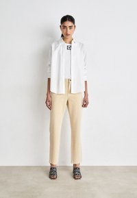 Camicia bianca a pulsante sopra una maglia nera con grafica, pantaloni beige e scarpe scivolate nere con logo audace. Design semplice e moderno.