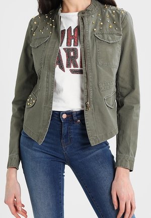 Woman wearing olive green studded military-style jacket over white graphic t-shirt and blue jeans, posing with hands relaxed.