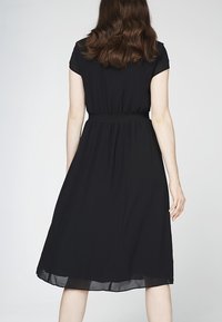 Schwarzes Kleid mit kurzen Ärmeln und elastischem Taillenbund. Der Stoff ist leicht, mit einem fließenden Rock, der unter das Knie fällt.