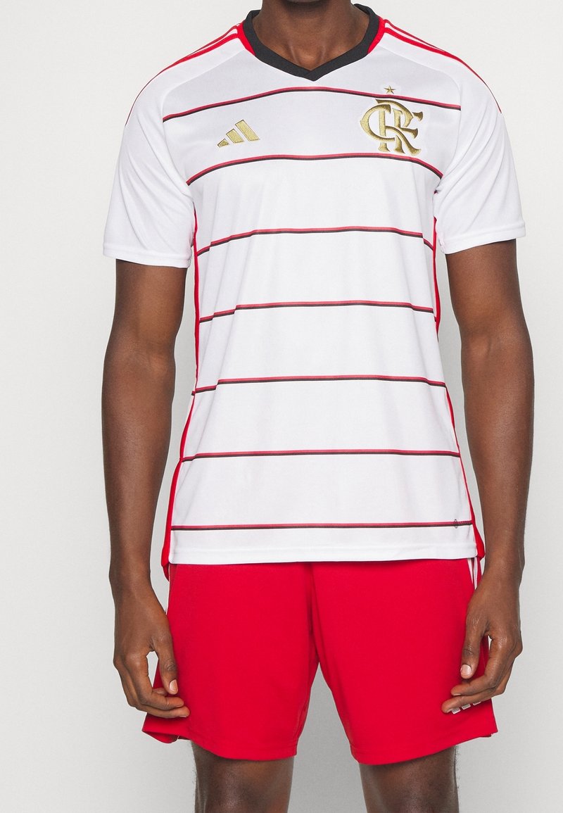 White short-sleeve soccer jersey with red stripes, black v-neck, and gold logo details. Red shorts complete the athletic outfit.