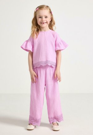 Lachend jong meisje met blond haar, gekleed in een roze top met ruches en korte mouwen en bijpassende wijde broek met kanten randje, staand binnen.