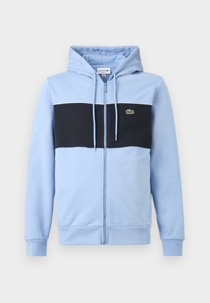 Hellblauer Kapuzenpullover mit Reißverschluss, mit schwarzem horizontalem Streifen über der Brust und kleinem grünem Lacoste-Logo auf der linken Seite.