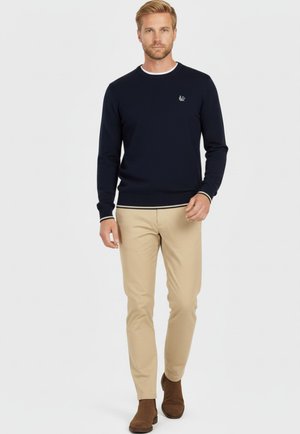 Suéter de cuello redondo en navy con un acento blanco en el collar, combinado con pantalones chinos beige y botas de tobillo marrones. Logo sutil en el lado izquierdo.