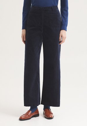 Persona con pantalones negros de pierna ancha, camiseta azul de manga larga, calcetines azules y mocasines de cuero marrón, de pie sobre un fondo blanco.