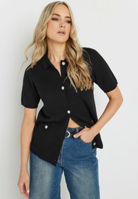 Svart kortärmad skjorta med krage, prydd med silverfärgade knappar och två framfickor. Mjuk textur, avslappnad passform, bärs med blå jeans.