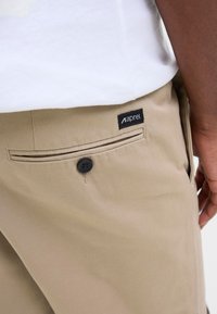 Poche de pantalon beige avec bouton noir et étiquette de marque en tissu noir, portée par une personne en chemise blanche, main reposant sur le côté.