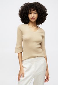 Beige gestricktes Oberteil mit einem runden Halsausschnitt und dreiviertellangen Glockenärmeln. Der Stoff wirkt weich und strukturiert und wird mit hellen Hosen kombiniert.