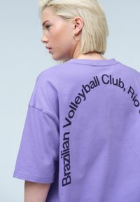 Ljubičasta oversized majica od mekog pamuka, s zakrivljenim crnim tekstom "Brazilian Volleyball Club, Rio" otisnutim na leđima.