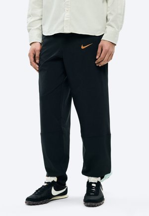Personne portant un pantalon Nike noir avec logo orange, un t-shirt blanc, des chaussettes blanches et des baskets Nike noires avec un swoosh blanc.
