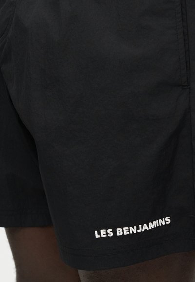 Μαύρο σορτσάκι με λευκό κέντημα "LES BENJAMINS" κοντά στο κάτω τελείωμα στο πόδι ενός ατόμου.