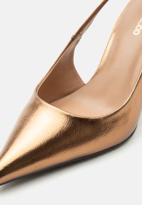 Κοντινή λήψη ενός μεταλλικού παπουτσιού με τακούνι σε απόχρωση μπρονζέ, με μυτερή μύτη, λεία και ελαφρώς υφασμένη επιφάνεια και εσωτερική επένδυση σε γυμνό χρώμα.