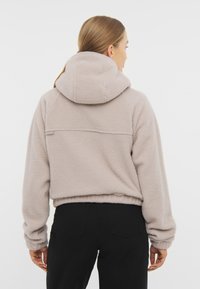 Dres w kolorze jasnobeżowym, wykonany z polaru, z wysokim kołnierzem i regulowanym kapturem, charakteryzujący się skróconym fasonem i miękkim, teksturowanym materiałem.