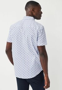 Camicia bianca a maniche corte con un motivo blu di piccoli forme ripetute. Presenta un colletto button-down e un orlo arrotondato. Realizzata in un tessuto leggero.