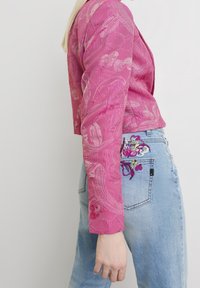 Veste rose texturée avec broderie florale sur les manches, dotée de deux boutons aux poignets, portée sur un jean bleu clair avec des accents floraux.