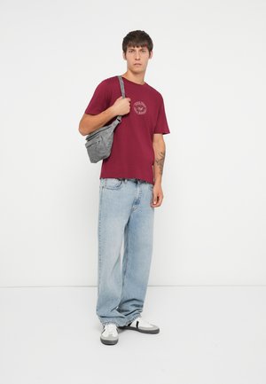 Burgundy cotton t-shirt with a round neckline and logo print, paired with light blue loose-fit jeans and gray crossbody bag. White sneakers.