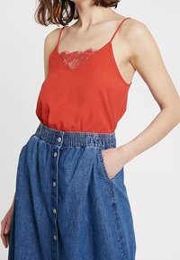 Camiseta sin mangas roja con detalles de encaje, combinada con una falda de mezclilla azul con cintura fruncida y botones en la parte delantera, manos en los bolsillos.