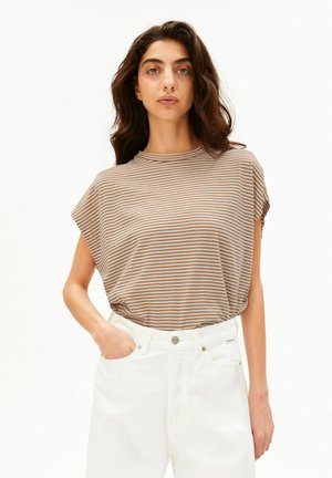 Donna con lunghi capelli scuri indossa una maglietta a maniche corte beige e bianco a righe infilata in pantaloni bianchi a vita alta, in piedi con una mano in tasca.