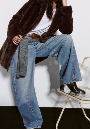 Personne portant un manteau en fourrure marron, une chemise blanche, un jean bleu à jambes larges et des bottes marron foncé, posant avec un pied sur une chaise transparente.