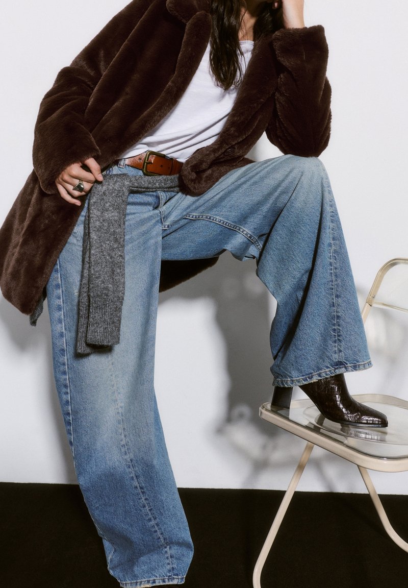 Personne portant un manteau en fourrure marron, une chemise blanche, un jean bleu à jambes larges et des bottes marron foncé, posant avec un pied sur une chaise transparente.