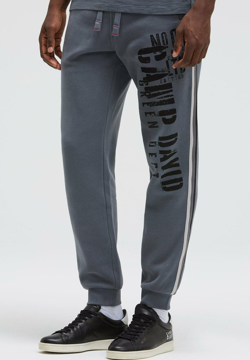 Graue Jogginghosen mit passgenauem Design, ausgestattet mit schwarzem Text-Branding an der Seite und grau-weißen Streifenakzenten entlang der Nähte.