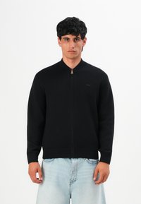 Jeune homme portant une veste zippée noire et un jean bleu clair, se tenant contre un fond blanc uni.