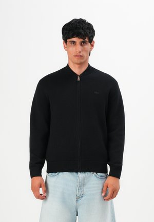 Jeune homme portant une veste zippée noire et un jean bleu clair, se tenant contre un fond blanc uni.