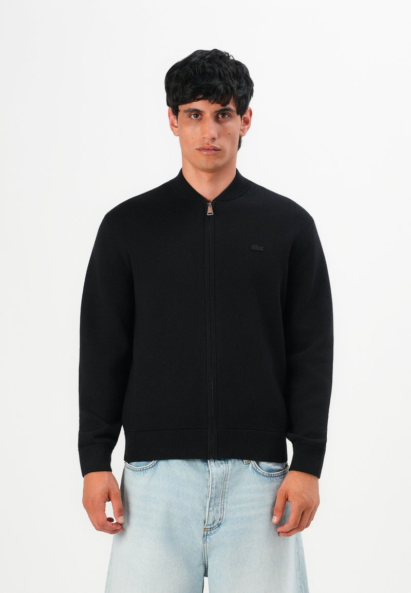 Jeune homme portant une veste zippée noire et un jean bleu clair, se tenant contre un fond blanc uni.