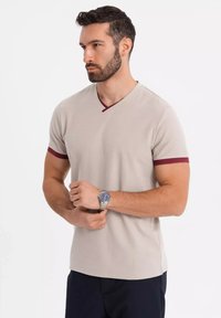 Beige V-hals t-shirt med vinrødt kantbånd på ærmer og halsudskæring. Korte ærmer, glat tekstur, iført et sølvur.