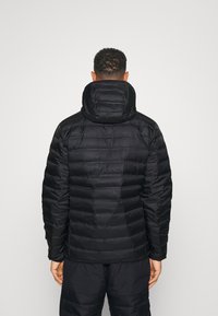Giacca nera imbottita con cappuccio, caratterizzata da sezioni trapuntate, una texture liscia e polsini elastici. Indossata sopra pantaloni neri coordinati.
