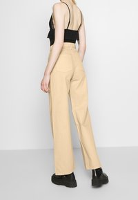 Pantalones de pierna ancha color beige, confeccionados en algodón, con una cintura alta, bolsillos traseros y un ajuste relajado. Combinados con un top corto de encaje negro.