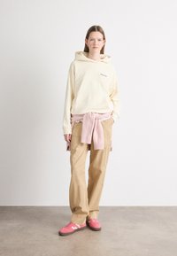 Sweat à capuche crème avec poche avant, pull rose noué à la taille, pantalon beige et baskets roses avec accents blancs. Fond uni.
