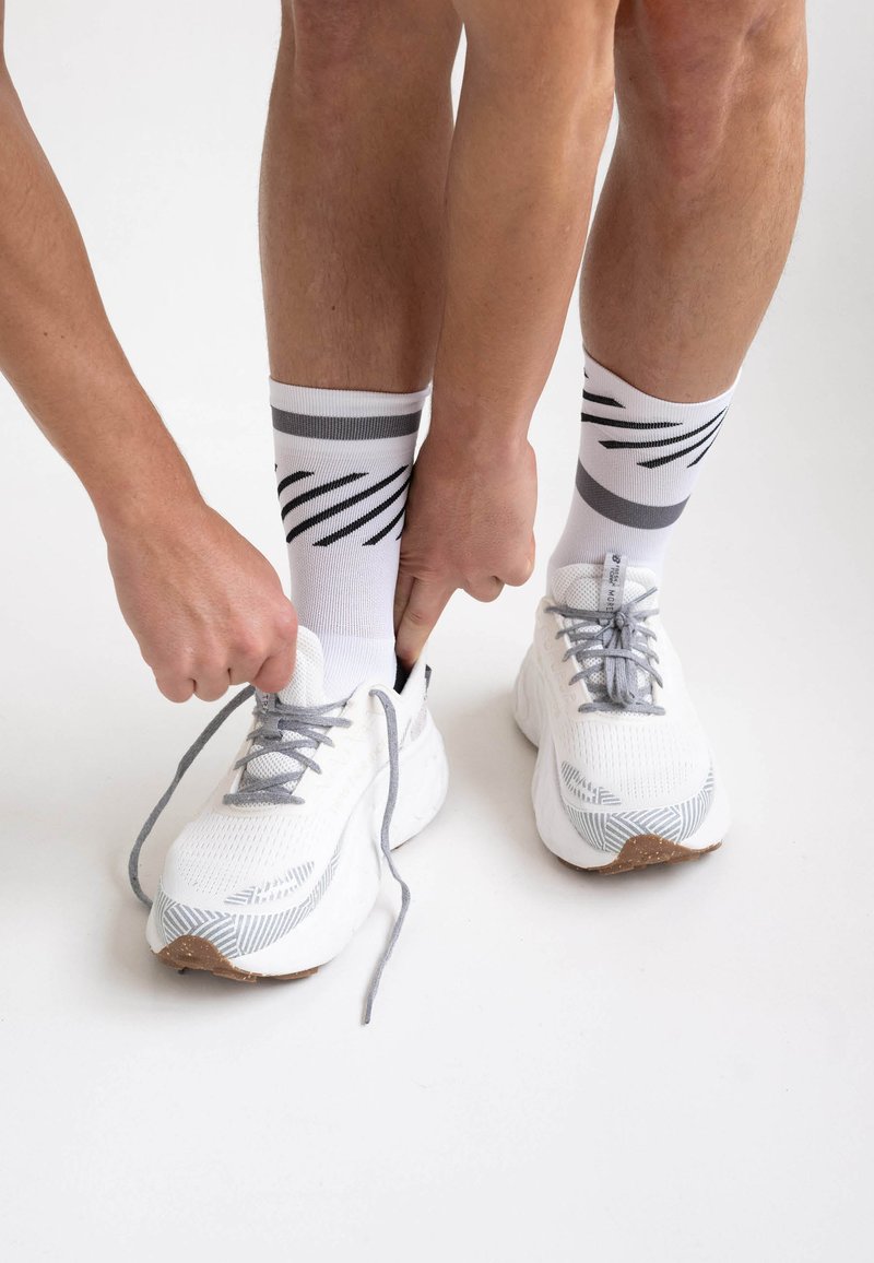 Weiße Sportschuhe mit grauen Akzenten und strukturierten Mustern, kombiniert mit weißen Socken mit schwarzen Streifen, die von zwei Händen geschnürt werden.