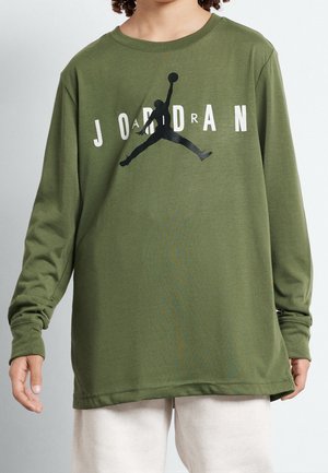 Person iført olivengrøn langærmet skjorte med sort Jumpman-logo og hvid "JORDAN"-tekst på brystet mod en ensfarvet baggrund.