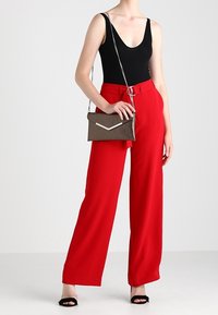 Pantalon rouge à jambes larges avec une taille ceinturée, associé à un body noir ajusté et une petite pochette marron avec des accents métalliques. Sandales à talons noires.
