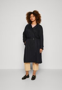 Marineblå trenchcoat med belte, dobbeltknappet design, lange ermer og sidelommer, kombinert med beige ankellange bukser og svarte loafers.