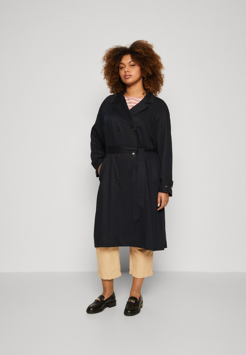 Marineblå trenchcoat med belte, dobbeltknappet design, lange ermer og sidelommer, kombinert med beige ankellange bukser og svarte loafers.