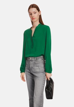 Grüne Bluse mit langen Ärmeln und V-Ausschnitt, kombiniert mit grauen hochgeschnittenen Jeans mit Fronttaschen und einer schwarzen Handtasche.