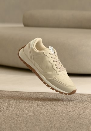Beige suède sneaker met lichtbruine rubberen zool, hangend boven een gestructureerd beige tapijt en neutrale bank op de achtergrond.