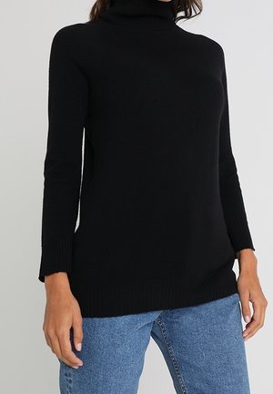 Femme portant un pull noir à col roulé et à manches longues, associé à un jean denim bleu sur un fond uni.